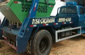 imagem de um caminhão poliguindaste azul, com uma caçamba verde sobre ele, estacionado em um galpão, com piso de terra